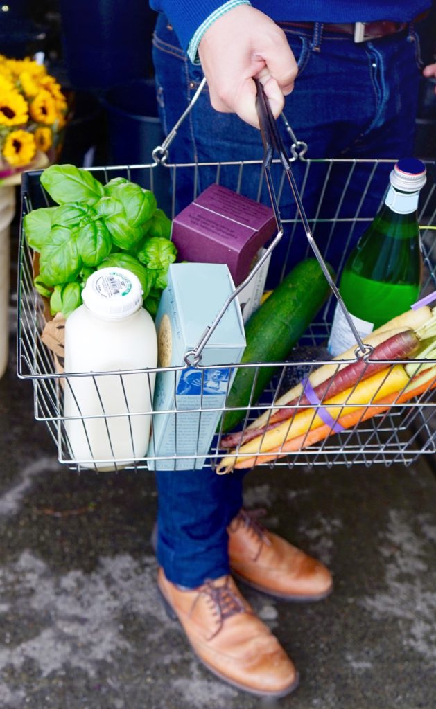 Einkaufskorb mit Basilikum, Milch, Mineralwasser, Karotten, Gurke, verpackten Lebensmitteln. Man sieht die Hand und die Beine des Mannes der ihn trägt.